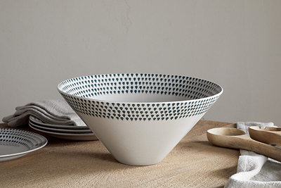 The nkuku Juniper Drop Serving Bowl - Cream & Juniper - Large features a wide rim and small dark blue dots around the top edge, making an inviting centrepiece on a beige textured surface with plates, folded napkins, and wooden utensils.
