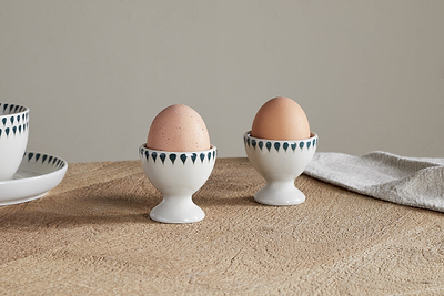 Two eggs are upright in nkuku’s Juniper Drop Egg Cups - Cream & Juniper (Set of 2), featuring a blue and green drop design, on a light wooden table with a beige cloth and a plate partly visible in the background.