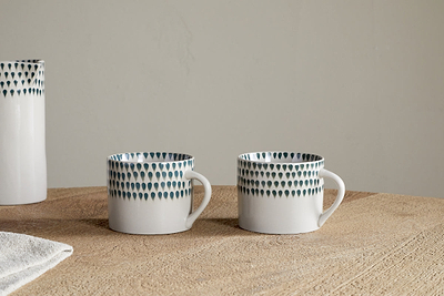 Two nkuku Juniper Drop Mugs in cream and juniper, featuring unique handles and signature drop design, rest side by side on a wooden surface. A matching vase and a white cloth are partially visible to the left. Set of 2 (Small).