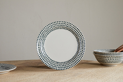 The nkuku Juniper Drop Side Plate in Cream & Juniper stands upright on a wooden surface, showcasing contemporary style. Matching bowls and a plate with a green drop pattern are partially visible nearby.