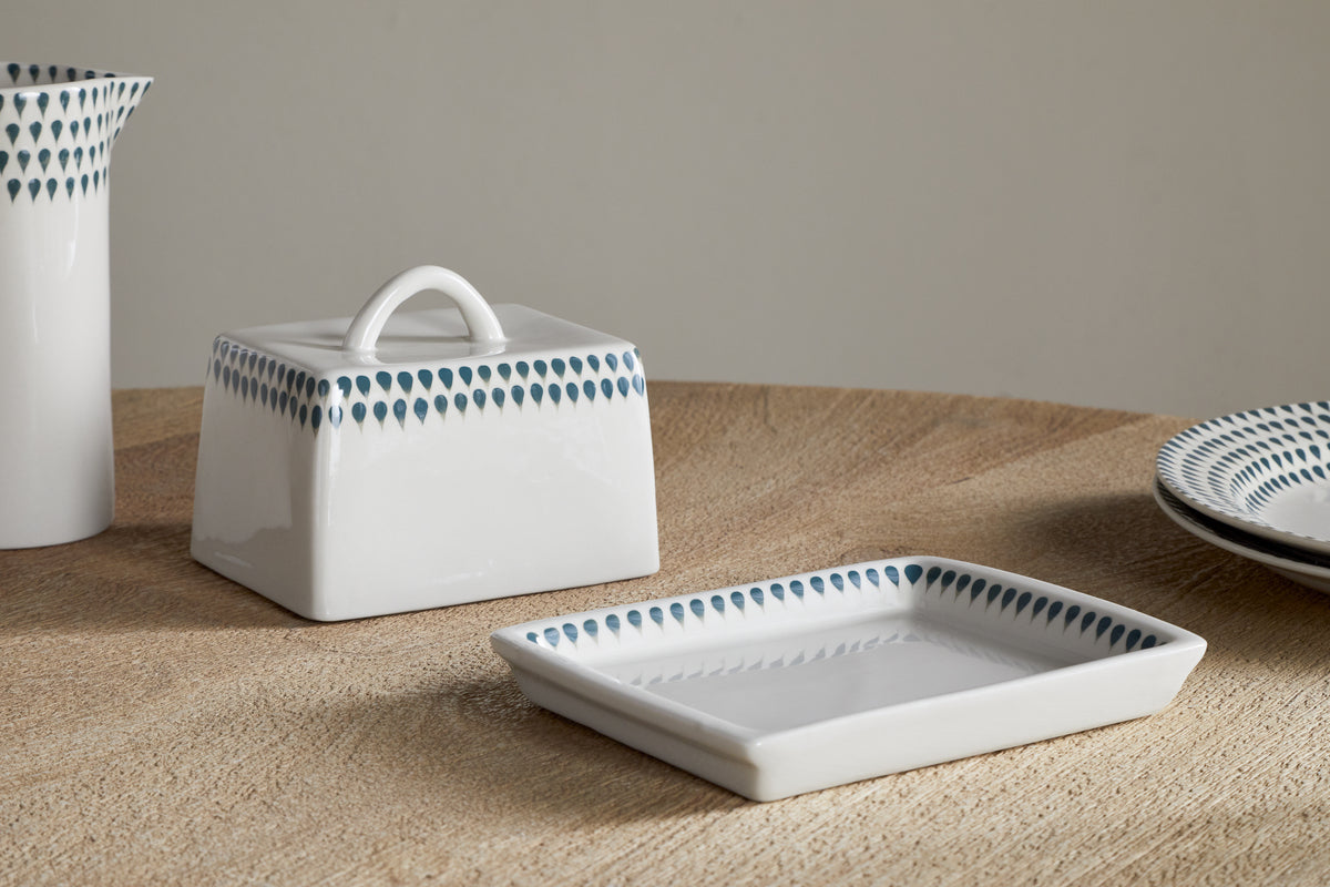 A close-up of nkukus Juniper Drop Butter Dish - Cream & Juniper, with a handle and blue teardrop edge pattern, sits alongside a matching rectangular plate on a wooden surface.