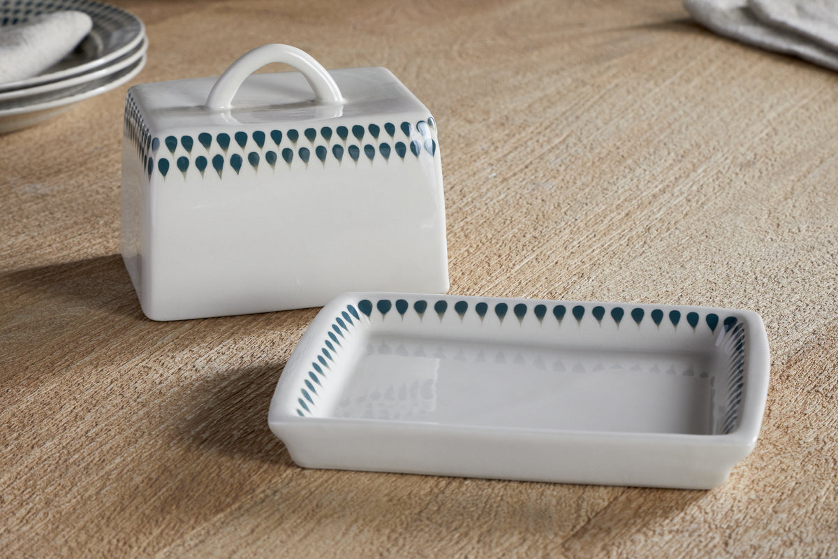 The nkuku Juniper Drop Butter Dish - Cream & Juniper, a rectangular ceramic dish with lid and blue-green drop design along the edges, is displayed on a wooden surface with the lid beside the base.