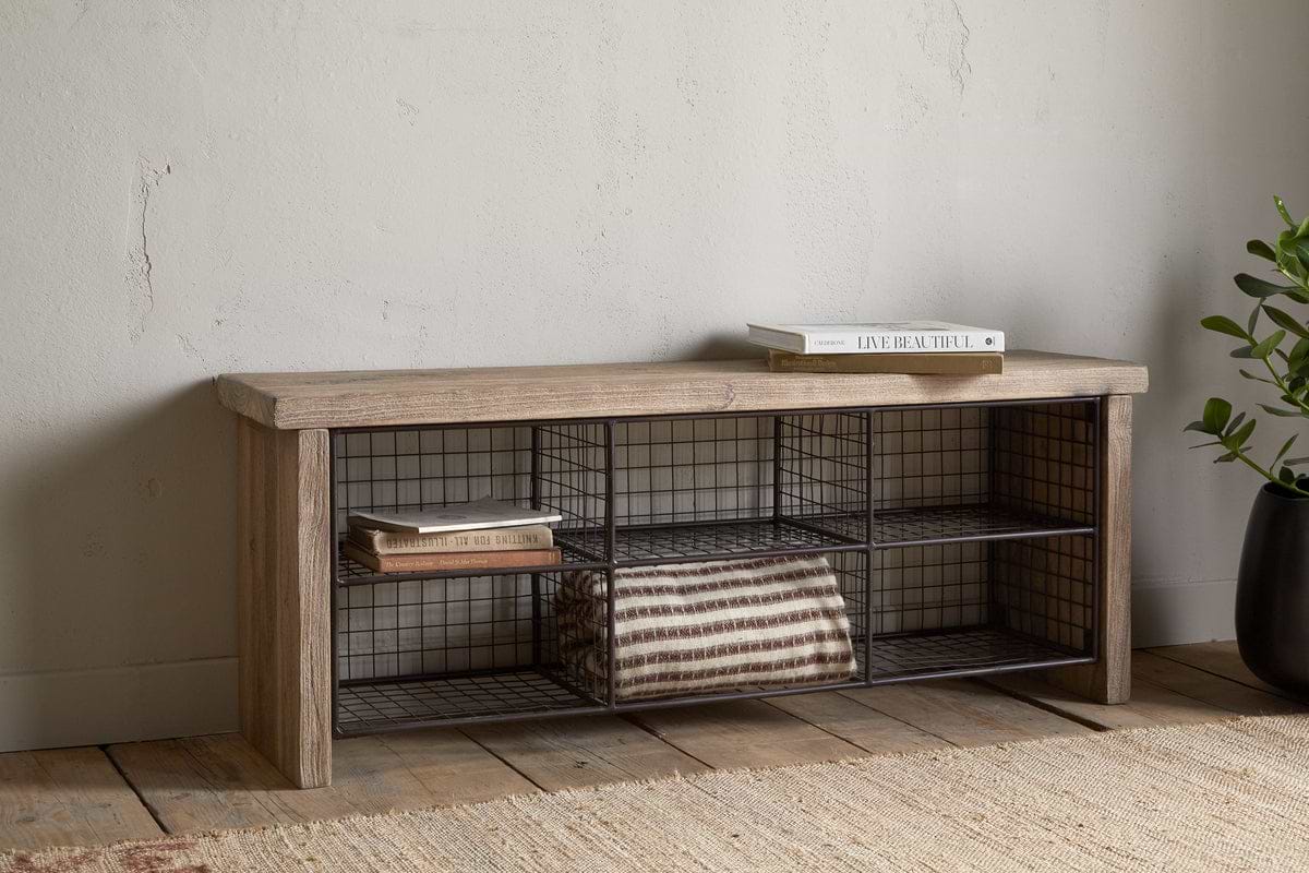 The nkuku Abha Reclaimed Wood & Iron Storage Bench - Natural holds a folded striped blanket and books, set against a pale wall by a potted plant on a woven rug—ideal as an entryway organizer.