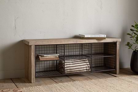 The nkuku Abha Reclaimed Wood & Iron Storage Bench - Natural holds a folded striped blanket and books, set against a pale wall by a potted plant on a woven rug—ideal as an entryway organizer.