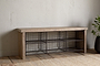 The nkuku Abha Reclaimed Wood & Iron Storage Bench – Natural is a versatile entryway organizer featuring six black wire baskets for shoe storage, set on a wooden floor beside a potted green plant.