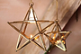 Two nkuku Antique Brass Glass Star t-light holders hang side by side, each with a glowing candle. Their gold frames and handmade glass catch the warm, blurred beige background light, highlighting the geometric lanterns.