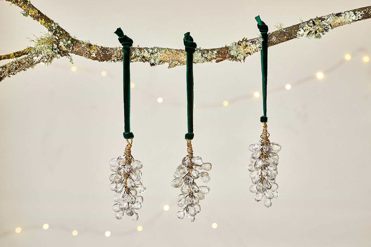 The Danvi Glass Drop Baubles - Brass & Clear (Set of 3) by nkuku hang from a tree branch, accented by twinkling string lights for a festive and elegant Christmas display.