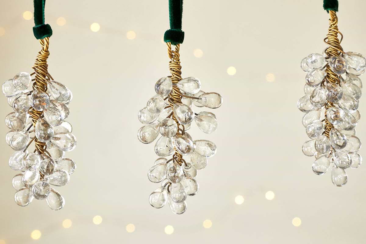 The nkuku Danvi Glass Drop Baubles - Brass & Clear (Set of 3) feature clusters of clear glass beads wrapped in brass wire and hang from a velvet tie, glowing with warm lights for a festive Christmas tree ambiance.