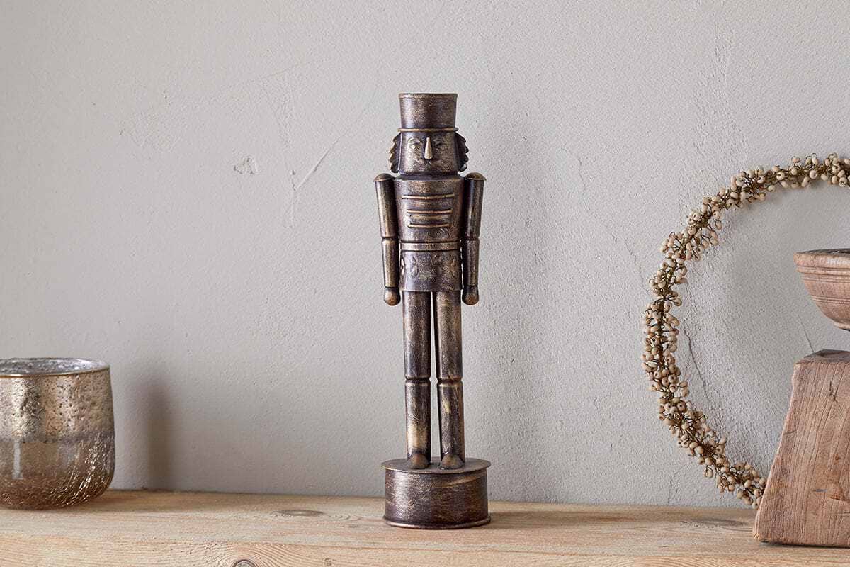 The nkuku Imay Nutcracker in antique brass stands on a wooden surface, accompanied by a silver candle holder, wreath, and bowl—creating an inviting seasonal display against a light gray wall.