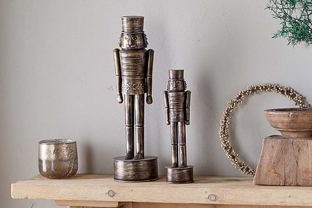 The Imay Nutcracker - Antique Brass by nkuku, in tall and short sizes, stands on a wooden shelf with a metallic candle holder, wooden bowl, and green foliage for a rustic, inviting seasonal display.