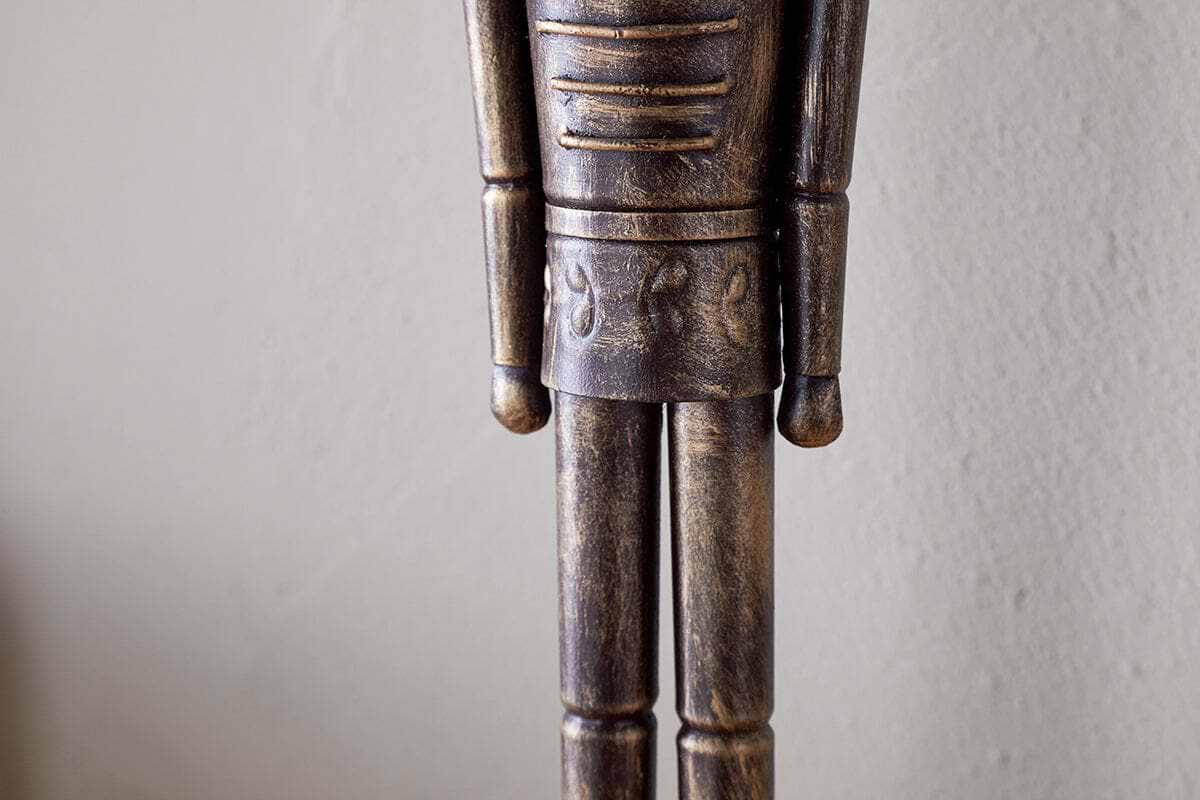 A close-up of the Imay Nutcracker - Antique Brass by nkuku shows its textured dark brown and gold finish, stylized cylindrical arms, and decorative waist carvings—perfect for festive or seasonal displays.