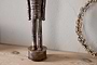 The Imay Nutcracker by nkuku, featuring an antique brass finish and elongated body, stands on a light wooden surface beside a dried floral wreath against a textured wall, creating a charming seasonal display.