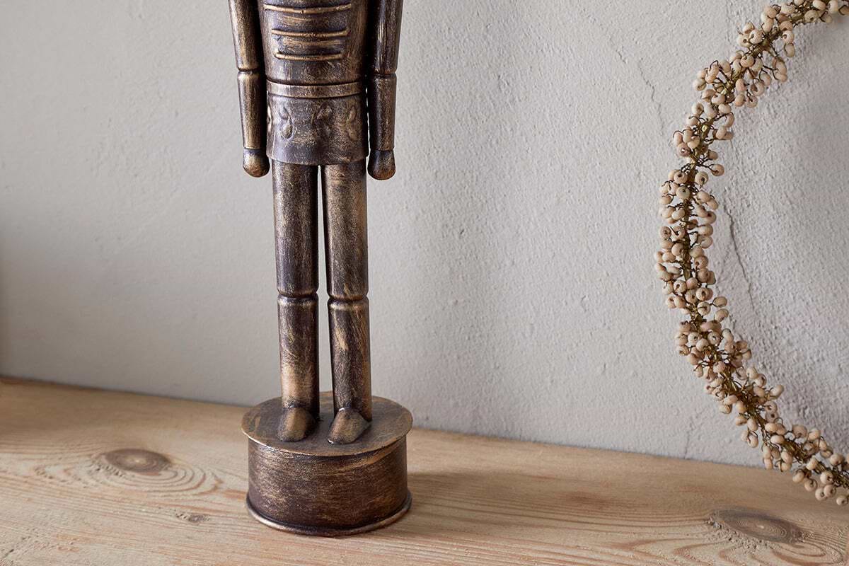 The Imay Nutcracker by nkuku, featuring an antique brass finish and elongated body, stands on a light wooden surface beside a dried floral wreath against a textured wall, creating a charming seasonal display.