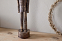 The Imay Nutcracker by nkuku, featuring an antique brass finish and elongated body, stands on a light wooden surface beside a dried floral wreath against a textured wall, creating a charming seasonal display.