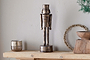 The nkuku Imay Nutcracker - Antique Brass stands on a mantel with a glass candle holder, round wreath, wooden bowl, and greenery against a light gray wall, creating an elegant seasonal display.