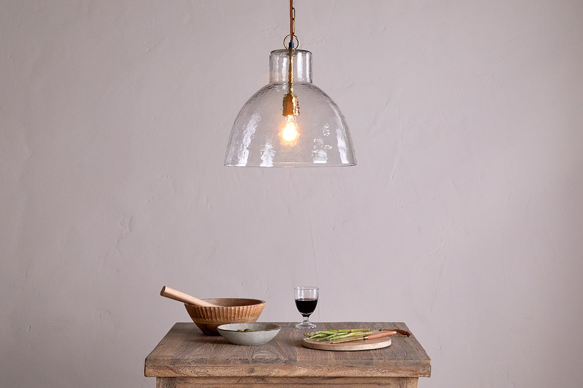 A wooden table displays a salad bowl, mortar and pestle, small gray bowl, cutting board with sliced veggies, and red wine. Above hangs the nkuku Inayah Recycled Glass Pendant - Clear with an exposed bulb against a plain beige wall.