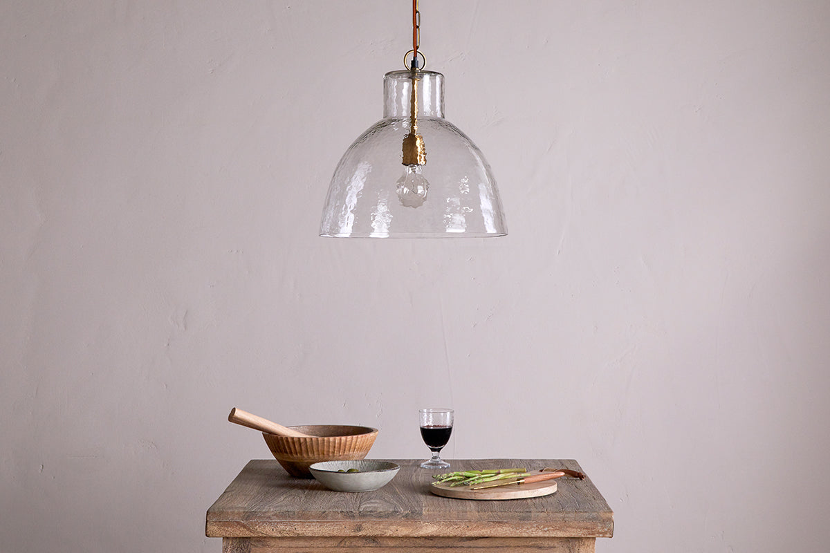 The Inayah Recycled Glass Pendant - Clear by nkuku hangs above a rustic wooden table set with a wooden bowl and pestle, gray bowl, glass of red wine, wooden board, knife, and sliced green vegetables against a plain light wall.