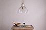 The Inayah Recycled Glass Pendant - Clear by nkuku hangs above a rustic wooden table set with a wooden bowl and pestle, gray bowl, glass of red wine, wooden board, knife, and sliced green vegetables against a plain light wall.