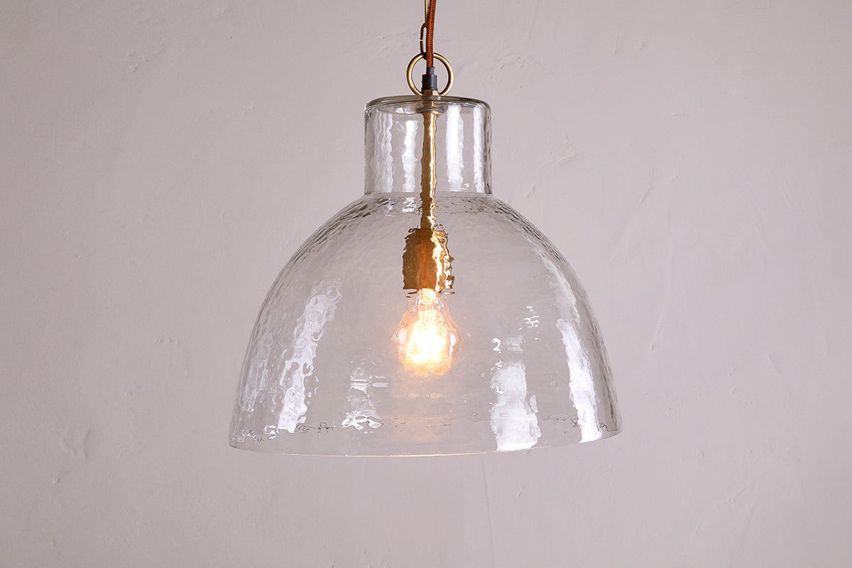 The Inayah Recycled Glass Pendant - Clear by nkuku features a dome-shaped, textured glass shade hanging from a gold fixture, illuminating the plain off-white wall with a warm, inviting glow.