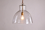 The Inayah Recycled Glass Pendant - Clear by nkuku features a dome-shaped, textured glass shade hanging from a gold fixture, illuminating the plain off-white wall with a warm, inviting glow.