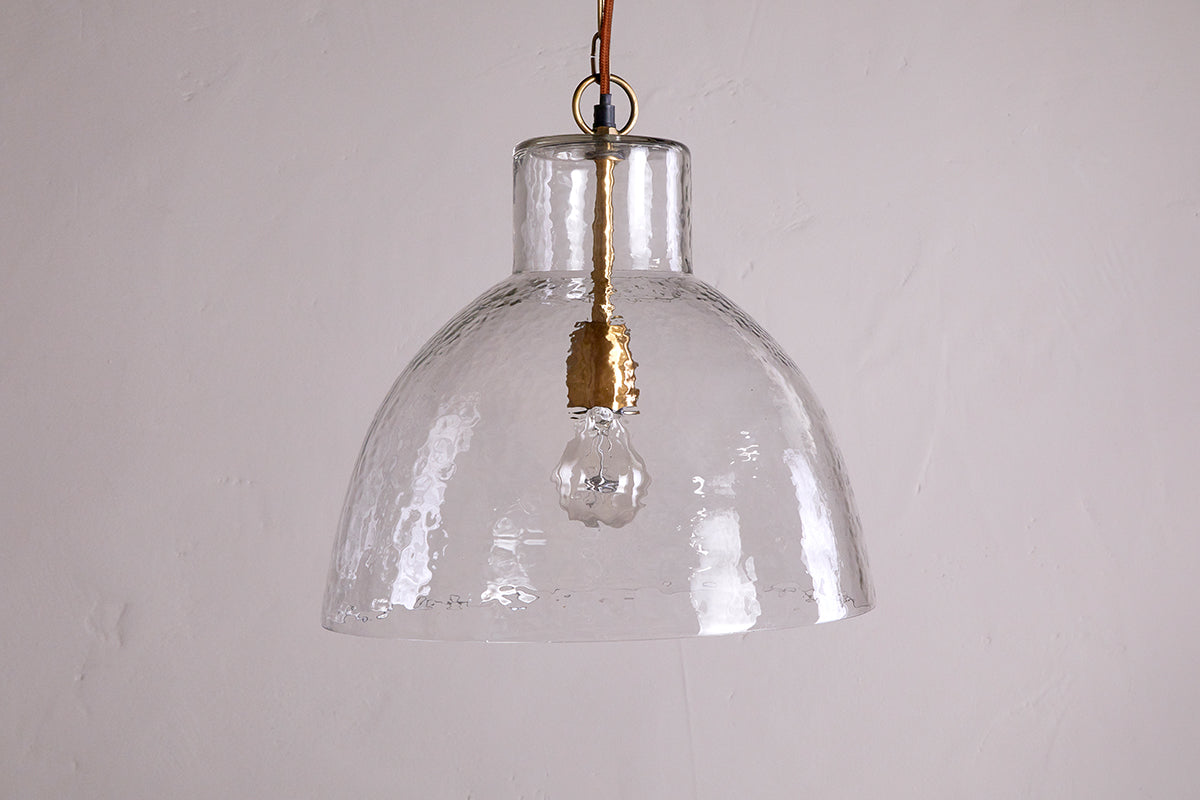 The Inayah Recycled Glass Pendant - Clear by nkuku features a wide, textured glass dome shade suspended by a chain, with a gold-toned socket visible inside, set against a plain, light background.