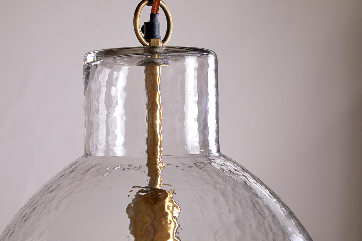 Close-up of the nkuku Inayah Recycled Glass Pendant in clear, featuring a textured glass shade, visible brass rod and fitting, and a brown cord connected to a round brass ring. The light hangs against a plain, light-colored background.