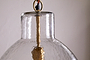 Close-up of the nkuku Inayah Recycled Glass Pendant in clear, featuring a textured glass shade, visible brass rod and fitting, and a brown cord connected to a round brass ring. The light hangs against a plain, light-colored background.