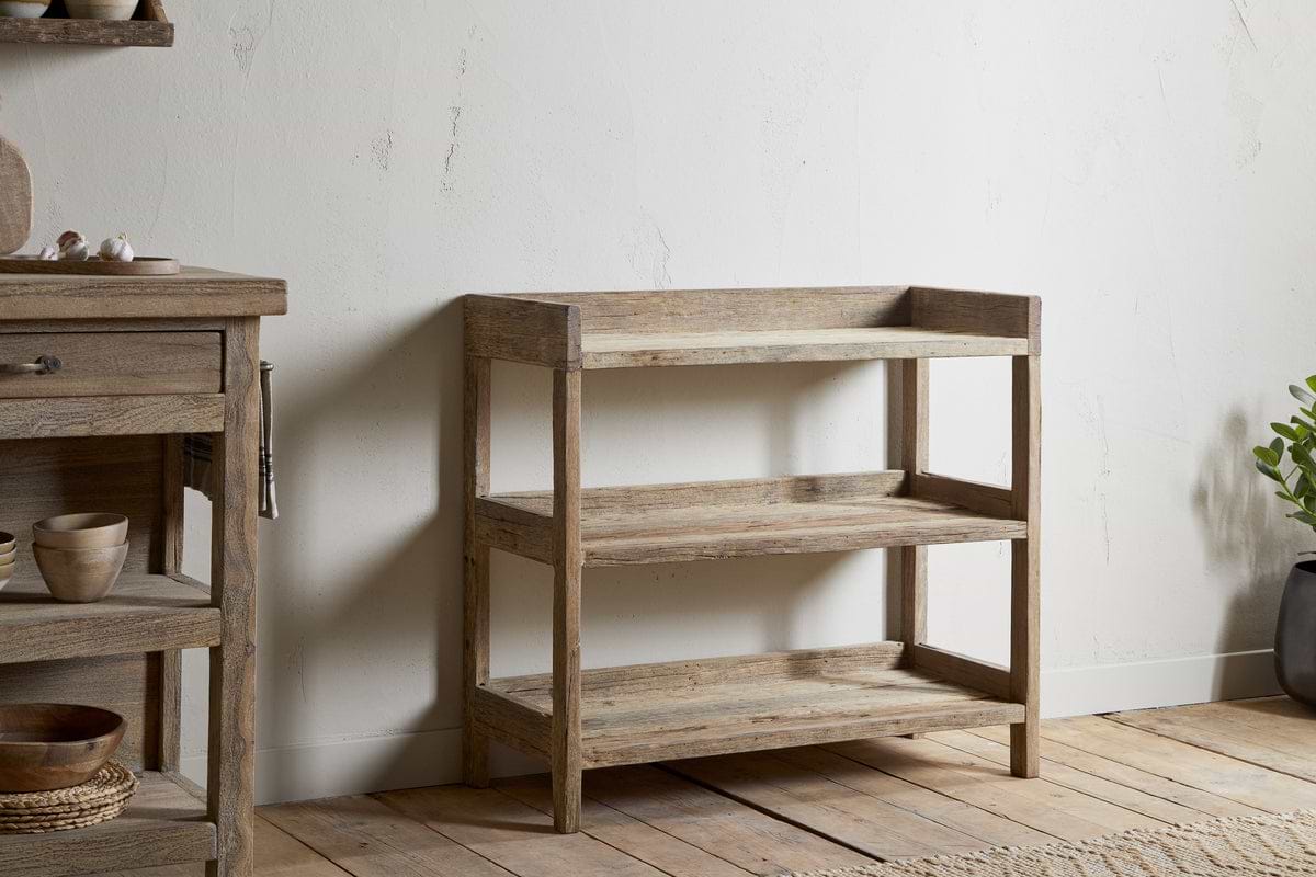 The nkuku Ibo Reclaimed Wood Pantry Standing Shelves in Natural, featuring a rustic three-tier open design, stands against a white textured wall on a wooden floor. A matching cabinet with bowls and a small potted plant complete the scene.