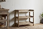 The nkuku Ibo Reclaimed Wood Pantry Standing Shelves in Natural, featuring a rustic three-tier open design, stands against a white textured wall on a wooden floor. A matching cabinet with bowls and a small potted plant complete the scene.