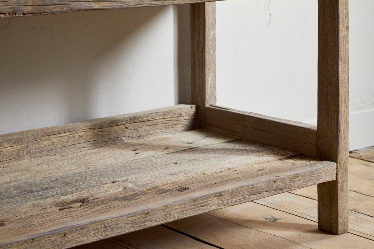 A close-up of the lower shelf and legs of the nkuku Ibo Reclaimed Wood Pantry Standing Shelves in Natural, resting on a light wooden floor against a white wall. The wood features a weathered, rustic finish.