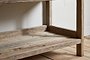 A close-up of the lower shelf and legs of the nkuku Ibo Reclaimed Wood Pantry Standing Shelves in Natural, resting on a light wooden floor against a white wall. The wood features a weathered, rustic finish.