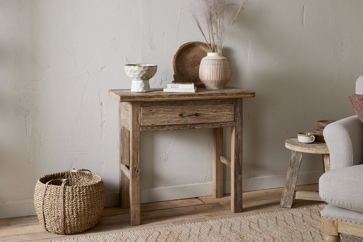 The nkuku Ibo Reclaimed Wood 1 Drawer Console Table in Natural brings rustic charm to your entryway, styled with a ceramic bowl, book, wooden tray, and vase with dried grasses. A woven basket sits nearby next to a cozy gray armchair.