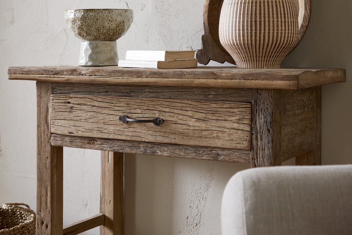 The Ibo Reclaimed Wood 1 Drawer Console Table by nkuku, styled with a textured drawer, ceramic bowl, two books, and a striped vase beside a beige chair and light wall, brings rustic charm to any entryway.