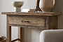 The Ibo Reclaimed Wood 1 Drawer Console Table by nkuku, styled with a textured drawer, ceramic bowl, two books, and a striped vase beside a beige chair and light wall, brings rustic charm to any entryway.