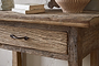 A close-up of the nkuku Ibo Reclaimed Wood 1 Drawer Console Table in Natural, showcasing a textured drawer with metal handle and weathered finish. Stacked books and a striped ceramic vase sit on top, set against a light entryway wall.