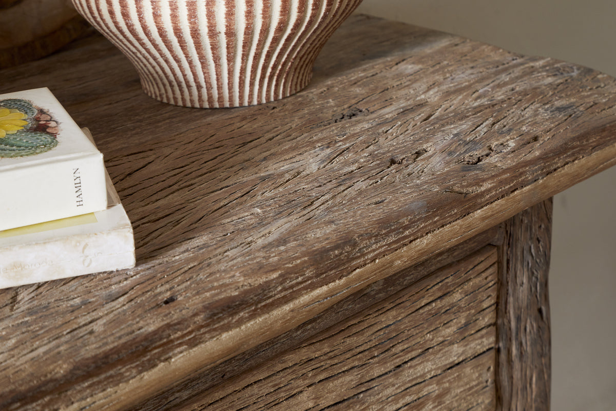 Close-up of the nkuku Ibo Reclaimed Wood 1 Drawer Console Table in Natural, its rustic surface displaying a striped ceramic vase and two stacked books, one with fruit illustrations on the cover.