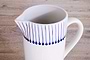 The nkuku Iba Jug - Indigo is a handmade white clay pitcher with a handle, vertical blue lines, dark blue dots near the rim, and a spout, shown on a light wooden surface.