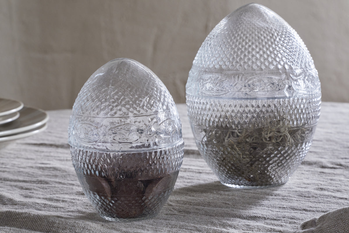 The nkuku Essan Recycled Glass Opening Eggs feature clear, textured recycled glass and decorative bands, making them stylish decor accents for any room. Their unique egg shape stands out beautifully on a beige fabric surface.