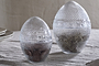 The nkuku Essan Recycled Glass Opening Eggs feature clear, textured recycled glass and decorative bands, making them stylish decor accents for any room. Their unique egg shape stands out beautifully on a beige fabric surface.