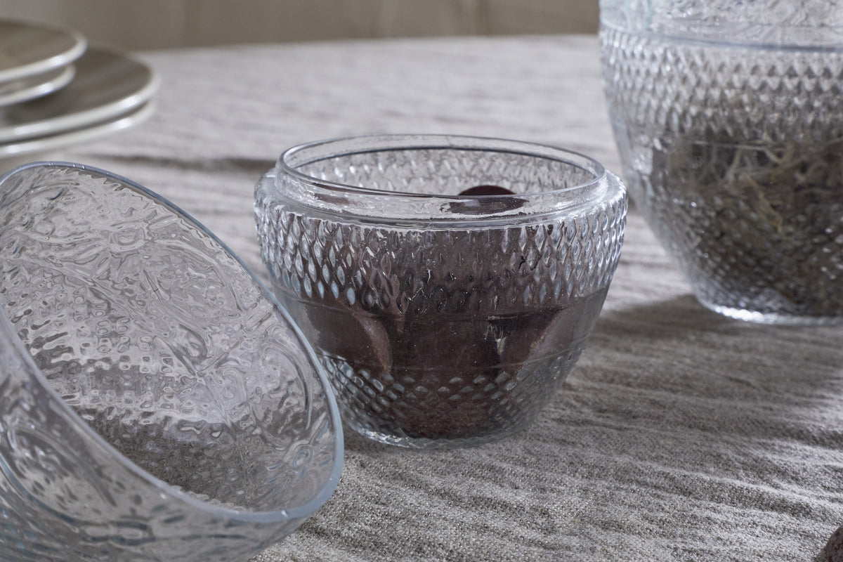 Three Essan Recycled Glass Opening Egg bowls by nkuku sit on a gray linen cloth—one open, another with chocolate and dark filling, the third with dried decor. Stacked plates blur in the background for a decorative touch.