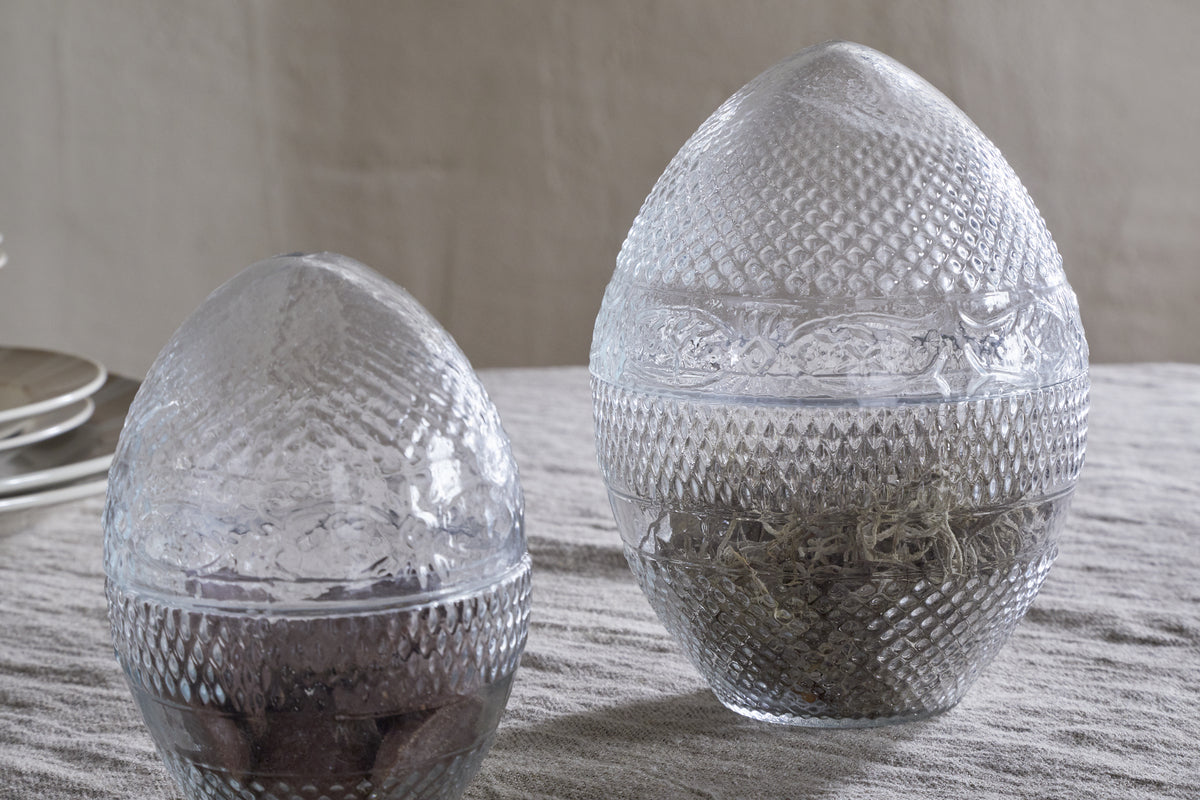 Two Essan Recycled Glass Opening Eggs by nkuku, crafted from clear recycled glass with lids, sit on beige fabric. Each egg contains natural materials—one with dried twigs, the other with a dark, coarse substance. Stacked plates appear behind them.