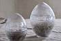 Two Essan Recycled Glass Opening Eggs by nkuku, crafted from clear recycled glass with lids, sit on beige fabric. Each egg contains natural materials—one with dried twigs, the other with a dark, coarse substance. Stacked plates appear behind them.