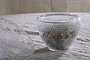 Two Essan Recycled Glass Opening Egg bowls by nkuku, with a diamond-patterned design, rest on a beige textured fabric—one upright holding dried plants, the other tipped on its side. Both are made from clear recycled glass.