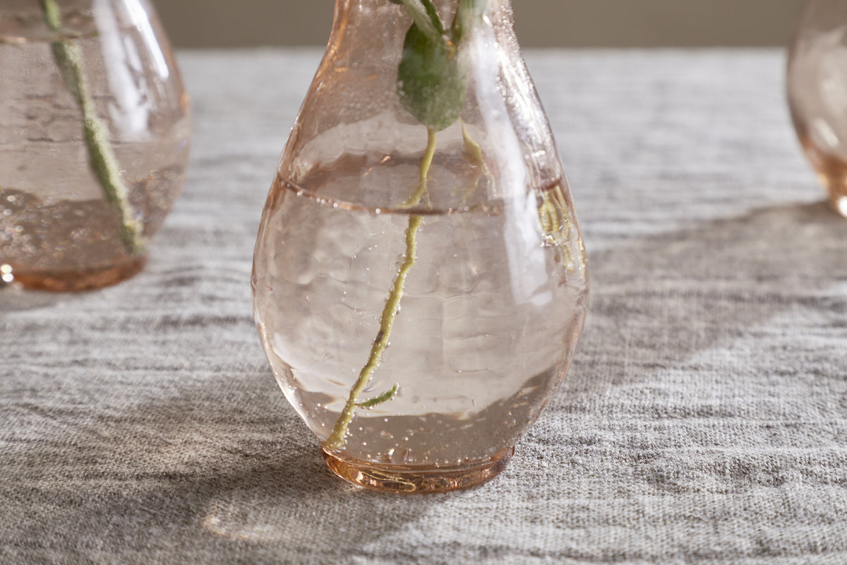 A set of Bapti Bud Vases in Dusty Terracotta by nkuku—three elegant glass vases with narrow necks, ideal for displaying plant stems. Shown on light gray fabric, they catch sunlight beautifully—perfect for lovers of stylish décor.