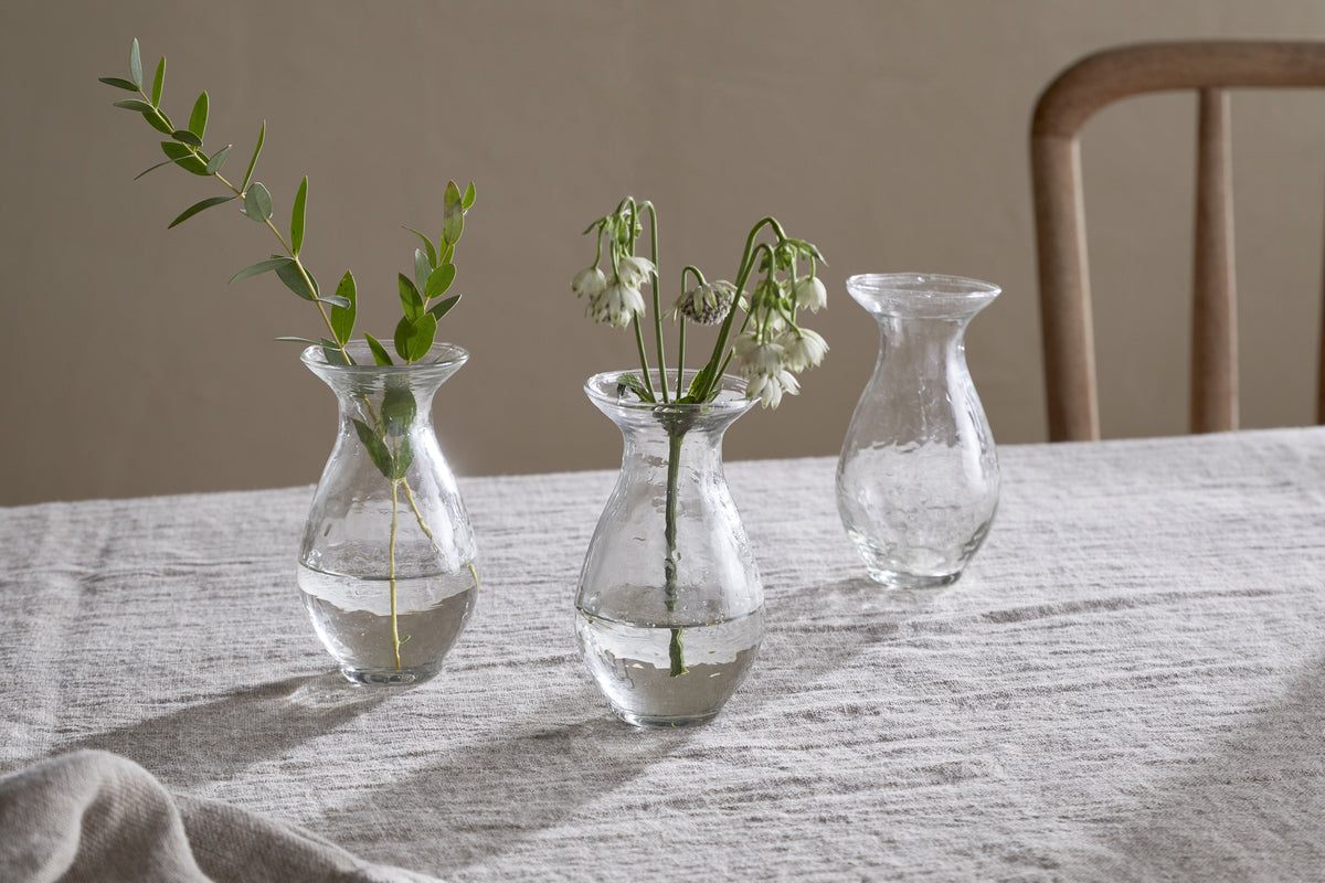 The nkuku Bapti Bud Vases - Clear (Set of 3) are displayed on a light gray linen tablecloth. One holds a leafy sprig, another has delicate white flowers, and the third is empty. A wooden chair can be seen in the background.
