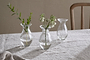 The nkuku Bapti Bud Vases - Clear (Set of 3) are displayed on a light gray linen tablecloth. One holds a leafy sprig, another has delicate white flowers, and the third is empty. A wooden chair can be seen in the background.