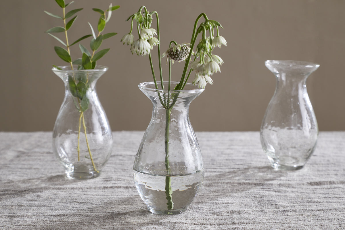 The Bapti Bud Vases (Set of 3) by nkuku sit on a light gray textured tablecloth. The left holds green foliage, the center has white drooping flowers, and the right is empty; all have water, rounded shapes, and flared rims.
