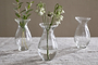 The Bapti Bud Vases (Set of 3) by nkuku sit on a light gray textured tablecloth. The left holds green foliage, the center has white drooping flowers, and the right is empty; all have water, rounded shapes, and flared rims.