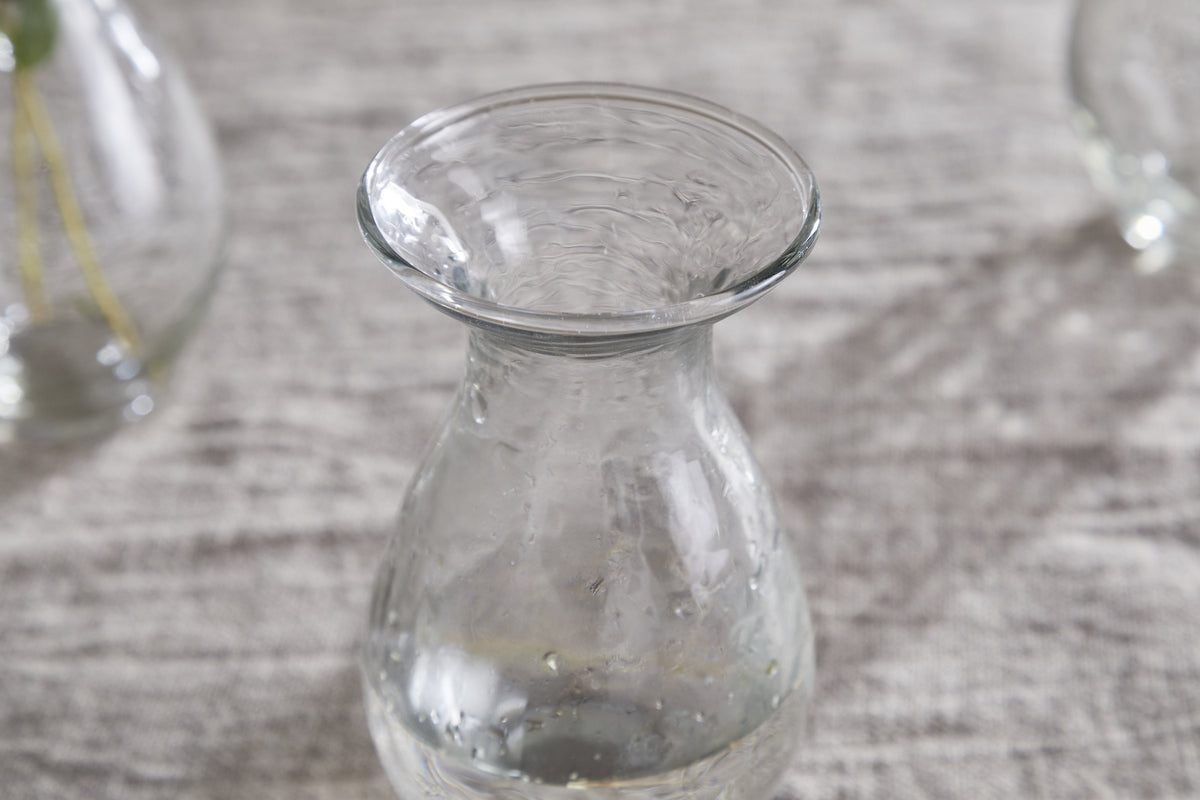 The nkuku Bapti Bud Vases - Clear (Set of 3) feature wide, flared rims and narrow necks. One vase with visible water droplets stands on a light gray textured surface, while two blurred vases are in the background.