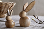 Two Khara Mango Wood Rabbit Standing Decorations by nkuku, featuring smooth rounded shapes and long ears, sit on a grey fabric surface. A wicker basket appears in the background left, with a budding branch to the right.